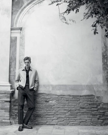 Portrait of a young adult male fashion model leaning against a brick and stucco wall in a beige jacket and black pants outdoors.