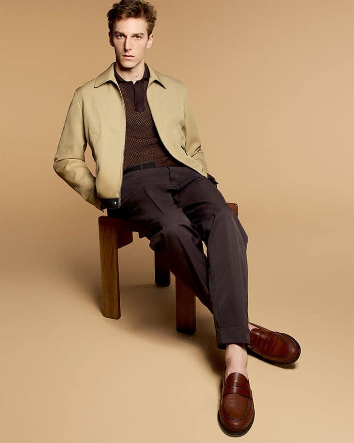 A fair-skinned adult man sits on a wooden stool in a studio setting wearing a tan jacket, brown polo shirt, and brown pants and shoes.