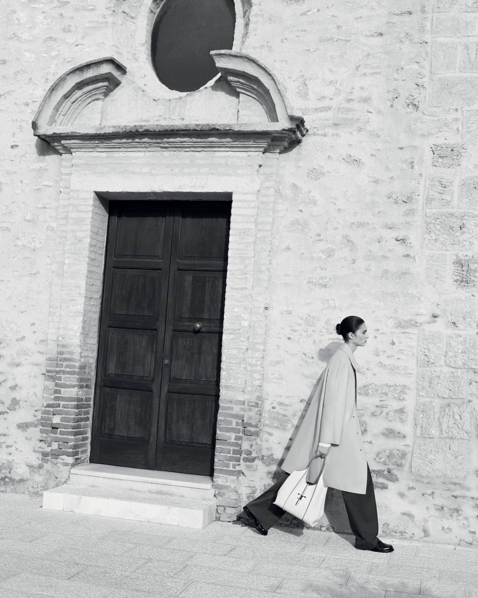Fashionable woman walks along a stone wall, wearing a tan coat, dark pants, and carrying a white tote bag; black and white.