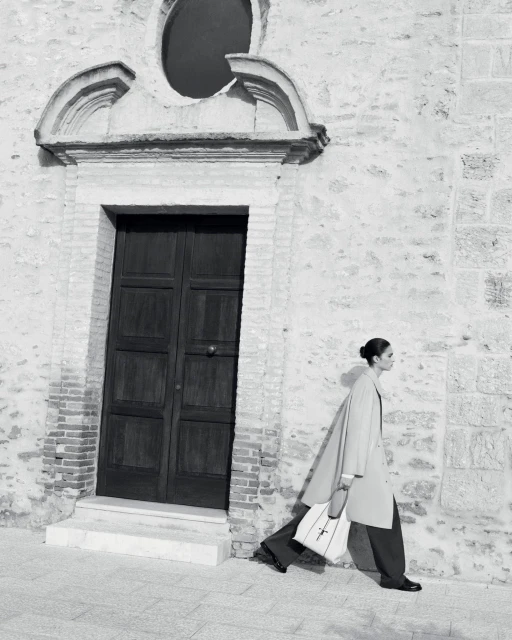 Fashionable woman walks along a stone wall, wearing a tan coat, dark pants, and carrying a white tote bag; black and white.