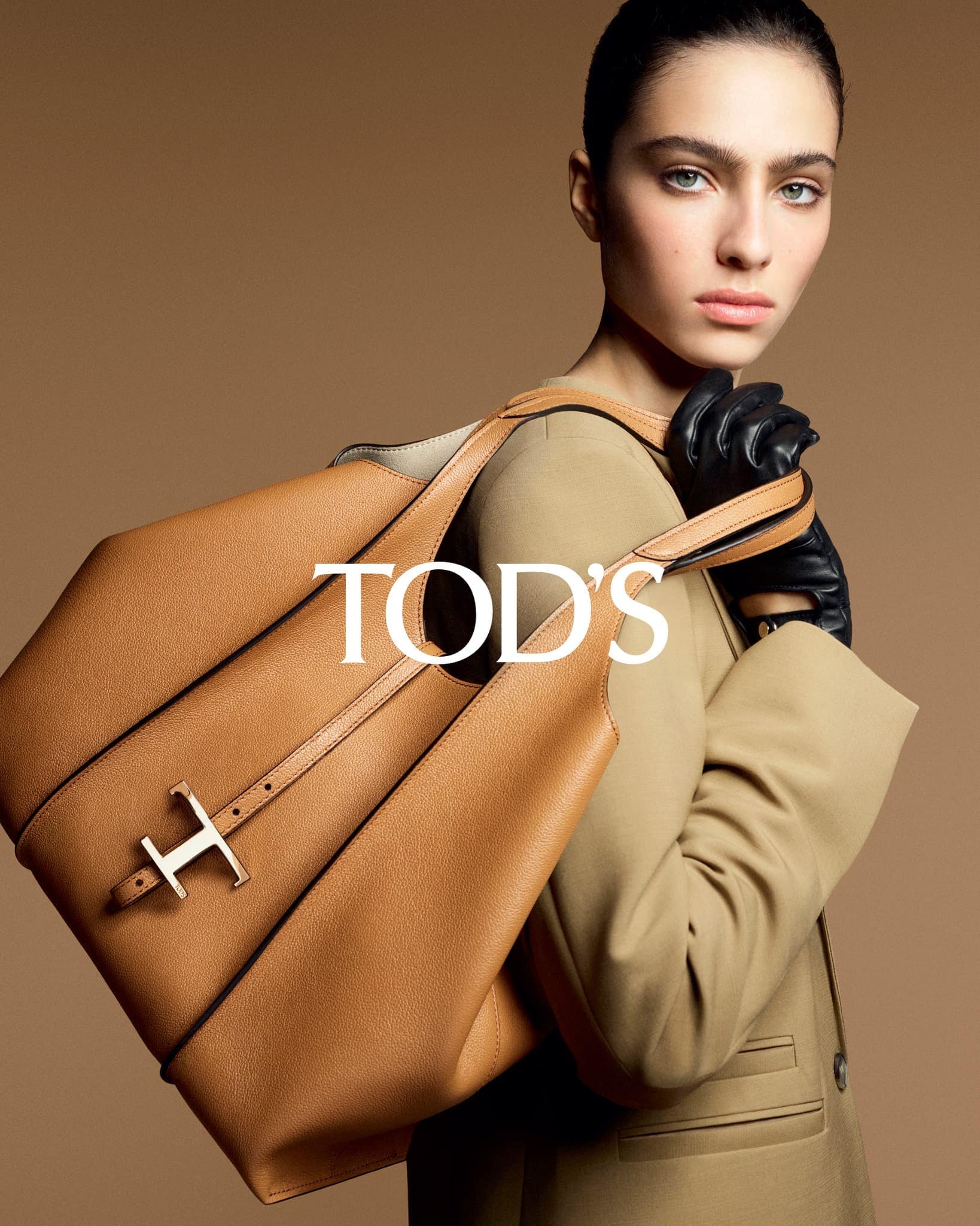 Fashion shot of a woman with dark hair and leather gloves holding a Tod's handbag in front of a neutral studio backdrop.