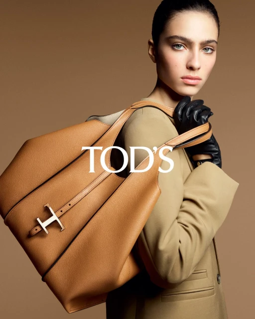 Fashion shot of a woman with dark hair and leather gloves holding a Tod's handbag in front of a neutral studio backdrop.