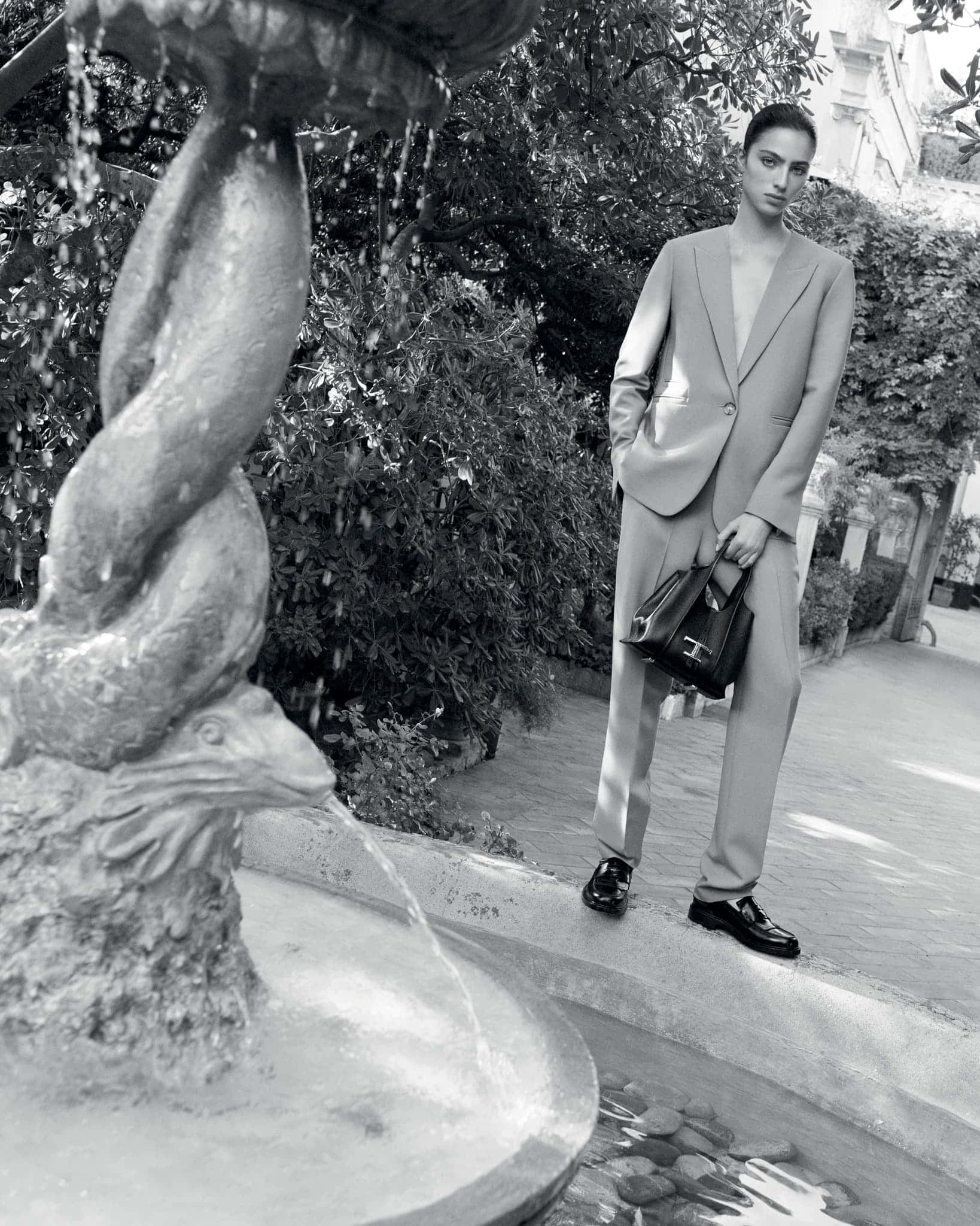 Black and white fashion shot: a woman in a suit holding a handbag stands next to a stone fountain in a garden setting with greenery.
