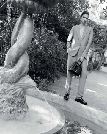 Black and white fashion shot: a woman in a suit holding a handbag stands next to a stone fountain in a garden setting with greenery.