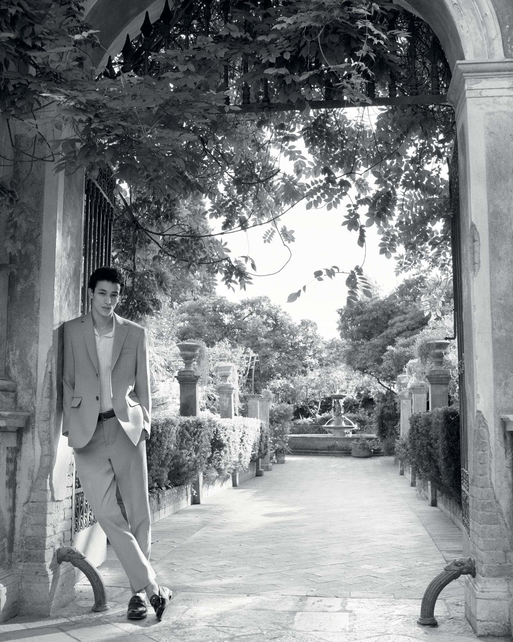 Man in a light-colored suit leans against an archway in a garden setting with trees, a fountain, and a stone-paved path in black and white.