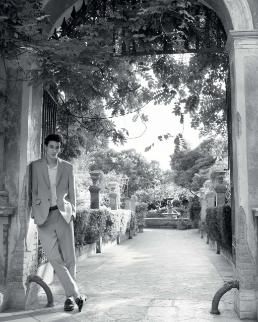 Man in a light-colored suit leans against an archway in a garden setting with trees, a fountain, and a stone-paved path in black and white.