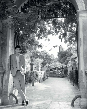 Man in a light-colored suit leans against an archway in a garden setting with trees, a fountain, and a stone-paved path in black and white.