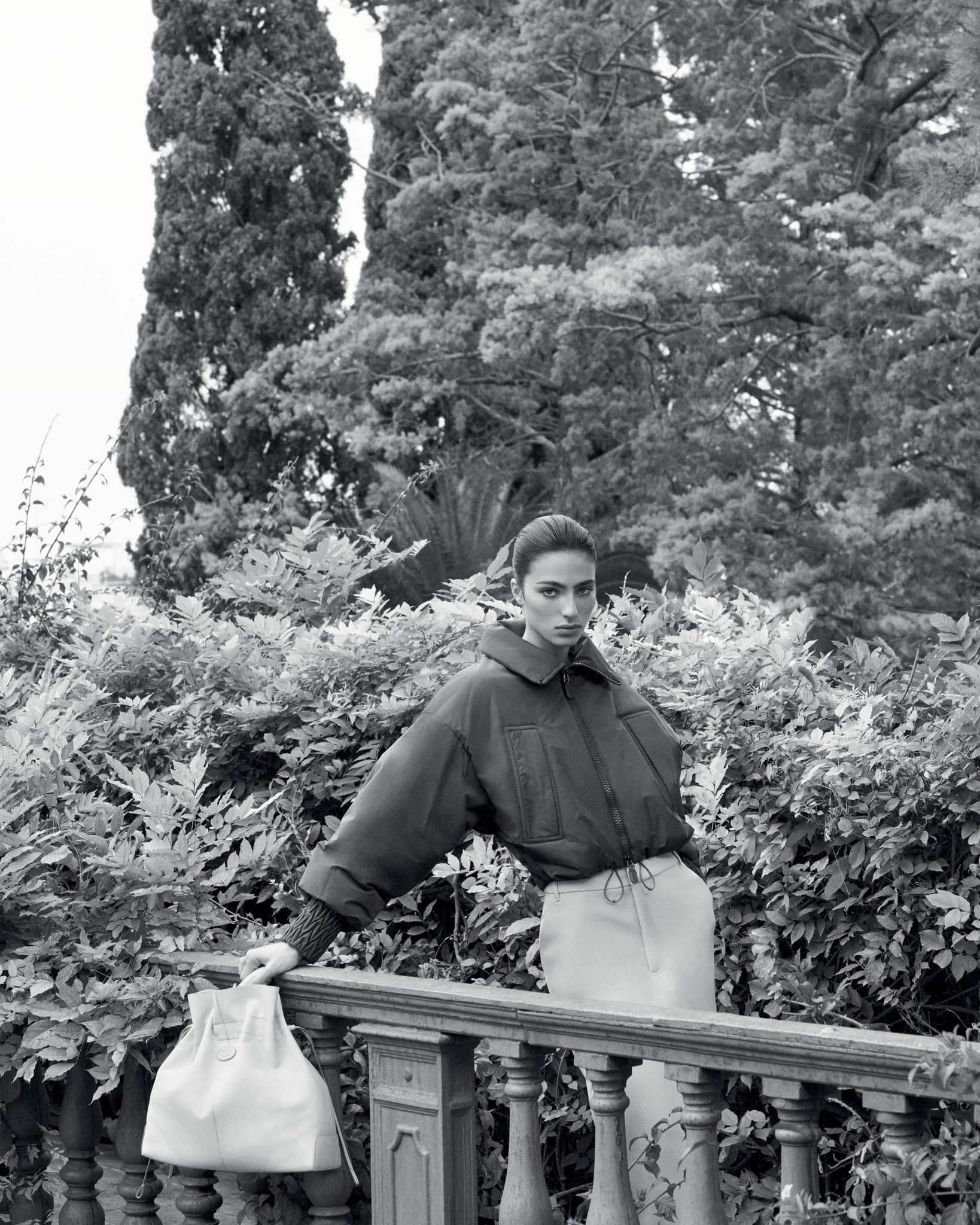 Fashion editorial shot of a woman in a puffy jacket posing outdoors near a railing and lush greenery, rendered in black and white.