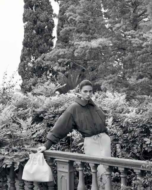 Fashion editorial shot of a woman in a puffy jacket posing outdoors near a railing and lush greenery, rendered in black and white.