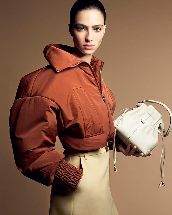 A fair-skinned woman poses in a brown puffer jacket and tan skirt, holding a white bucket bag in a studio shot on a neutral backdrop.