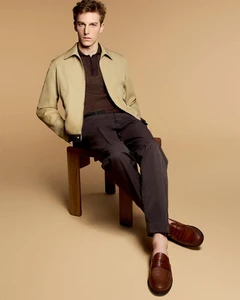 A young male model seated on a dark wooden stool wearing a beige jacket, brown polo, dark trousers, and leather loafers against a plain tan backdrop.