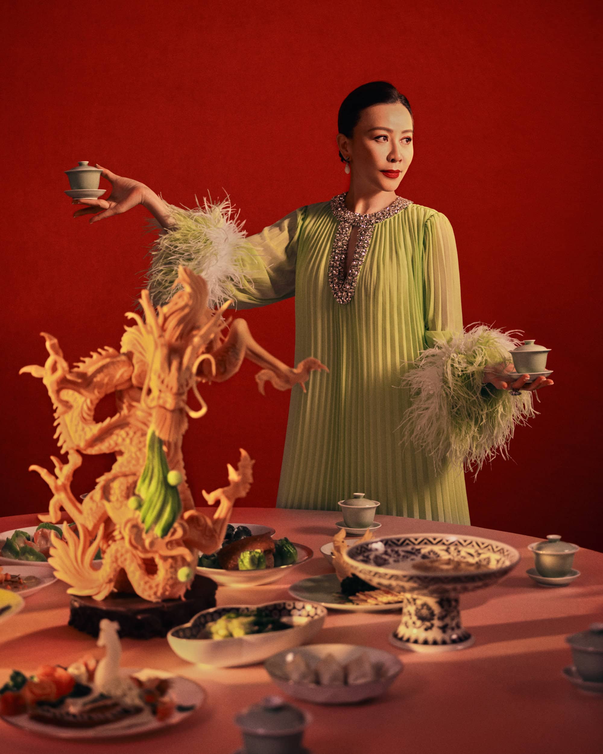 Adult female in a green dress stands behind a table with food and a decorative dragon against a red backdrop in a studio setting.
