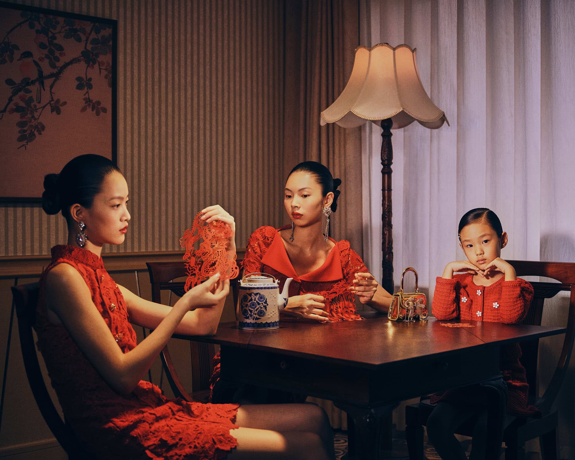 Three Asian females in red dresses sit at a table with red paper cut art, a teapot, and a lamp in an indoor setting with striped wallpaper.