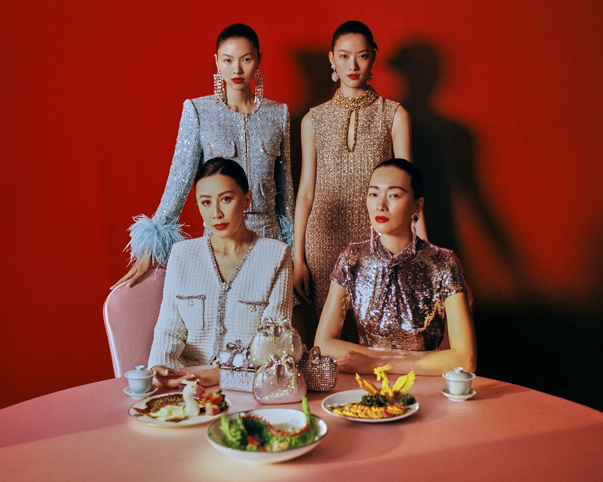 Four Asian women in fashionable dresses pose around a table set with dishes, teacups, and purses against a red background.