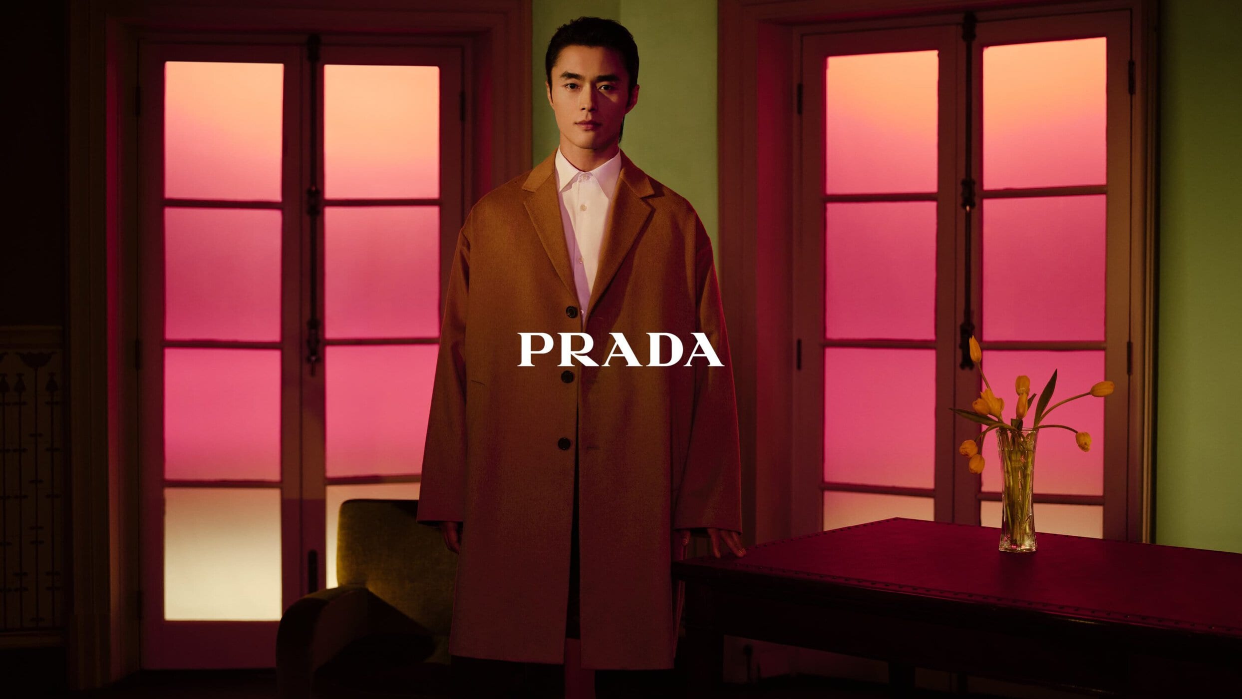 A stylish Asian male model showcases a long tan coat in a fashion-forward indoor setting, emphasizing modern elegance and brand appeal.