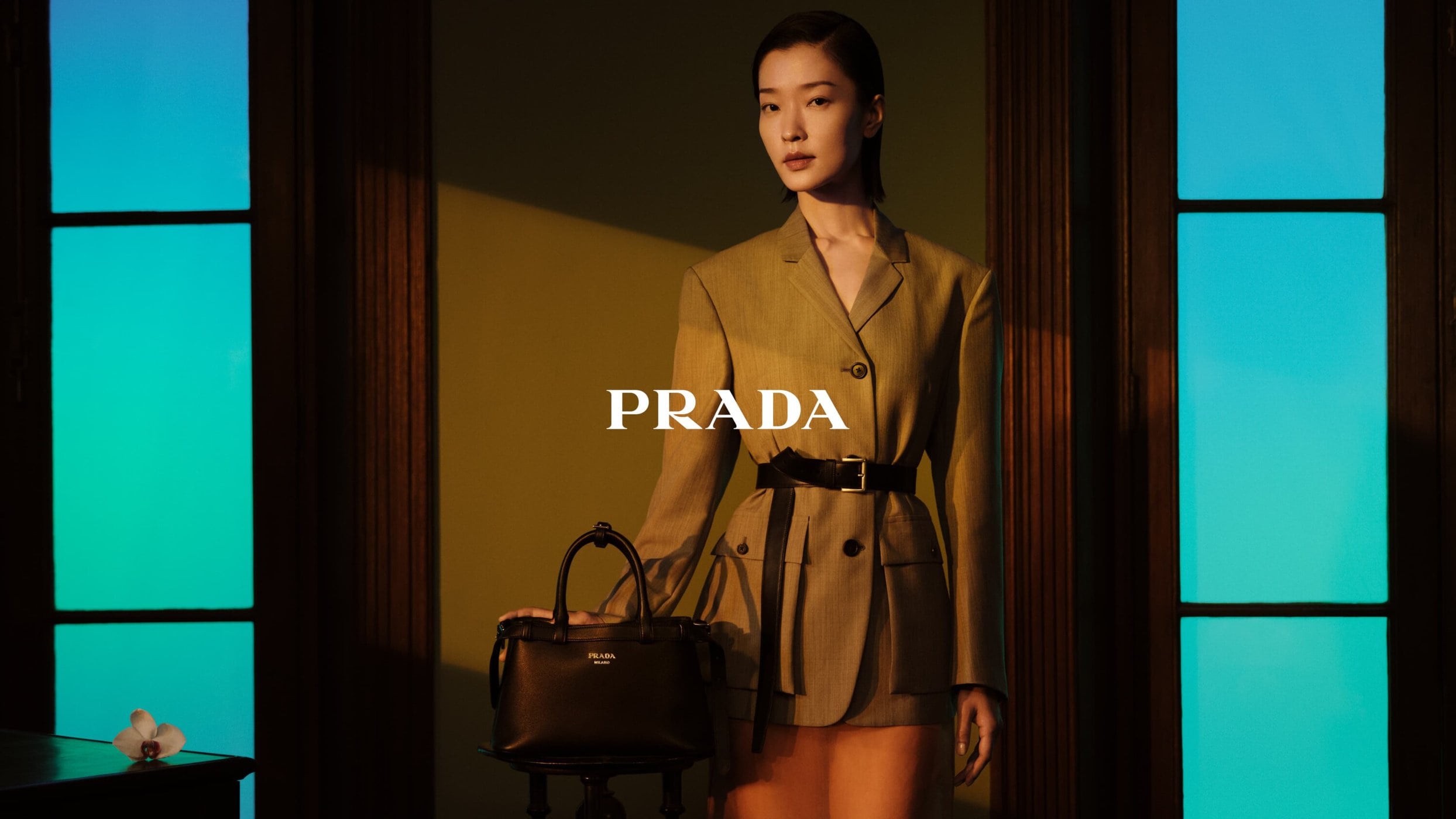 Asian woman in olive blazer dress holds black leather Prada handbag in studio with teal lighting. Fashion editorial shot.