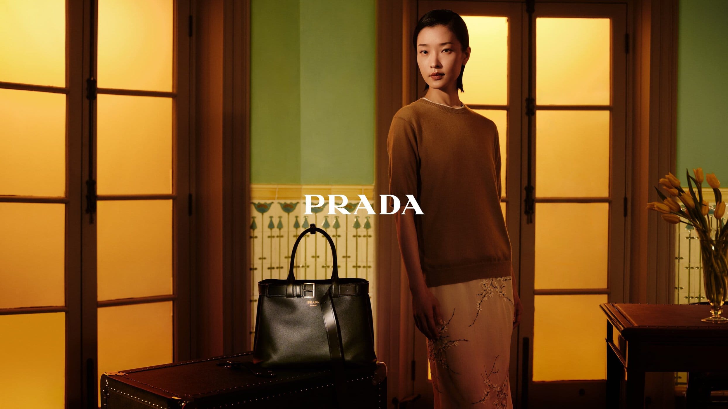 A fashionable Asian woman poses indoors with a Prada handbag, wearing a brown sweater and light skirt, lit by golden light.