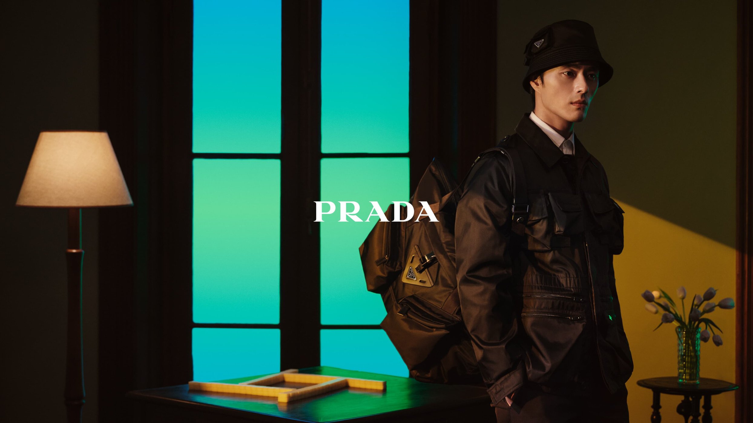 Stylish man poses indoors in Prada jacket and bucket hat, leaning on desk near vase of tulips, illuminated by vibrant gel lighting.