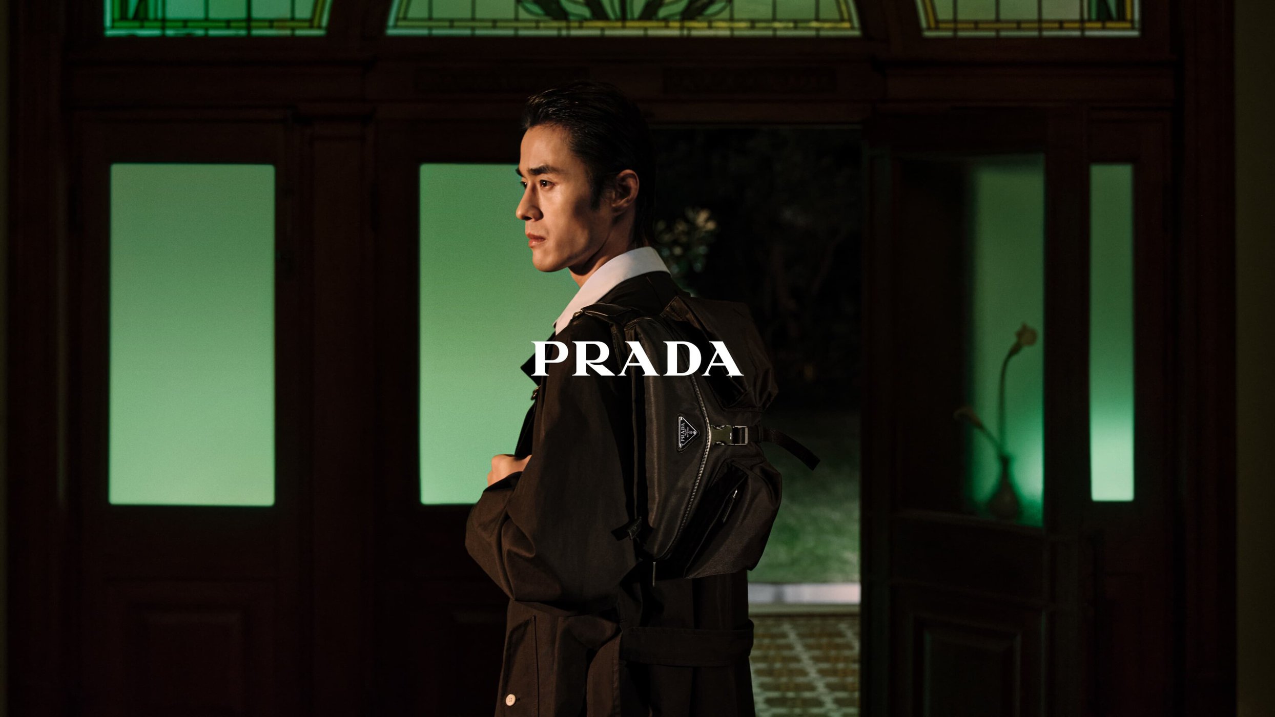 A side-profile shot of a stylish Asian man wearing a black trench coat and backpack, with the Prada logo prominently displayed.