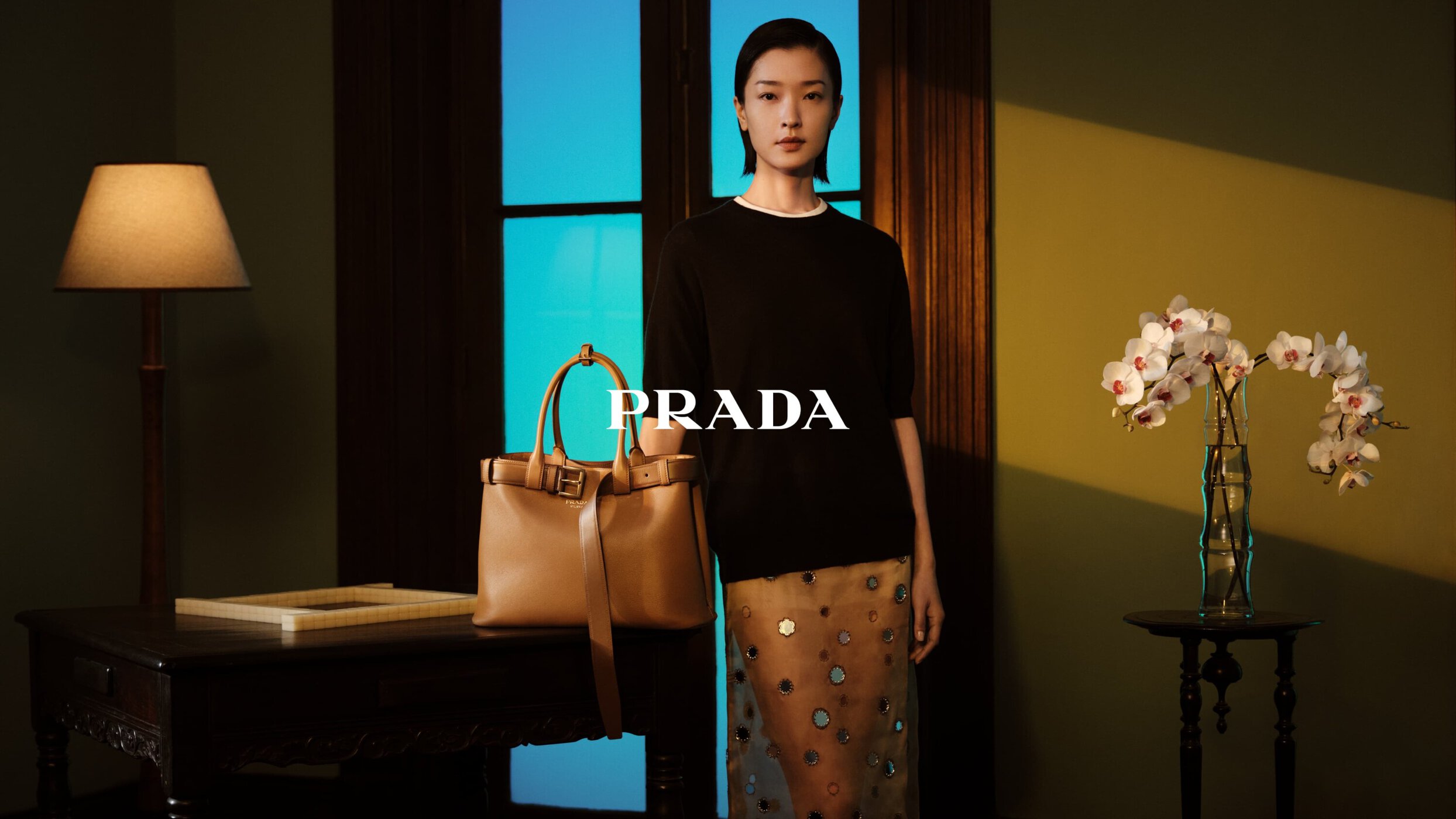 Asian woman poses indoors with a Prada handbag, wearing a black top and patterned skirt, next to a lamp, desk, and flower arrangement.