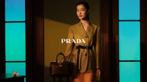 Model in a belted khaki blazer standing beside a black handbag inside an interior with glowing turquoise panels