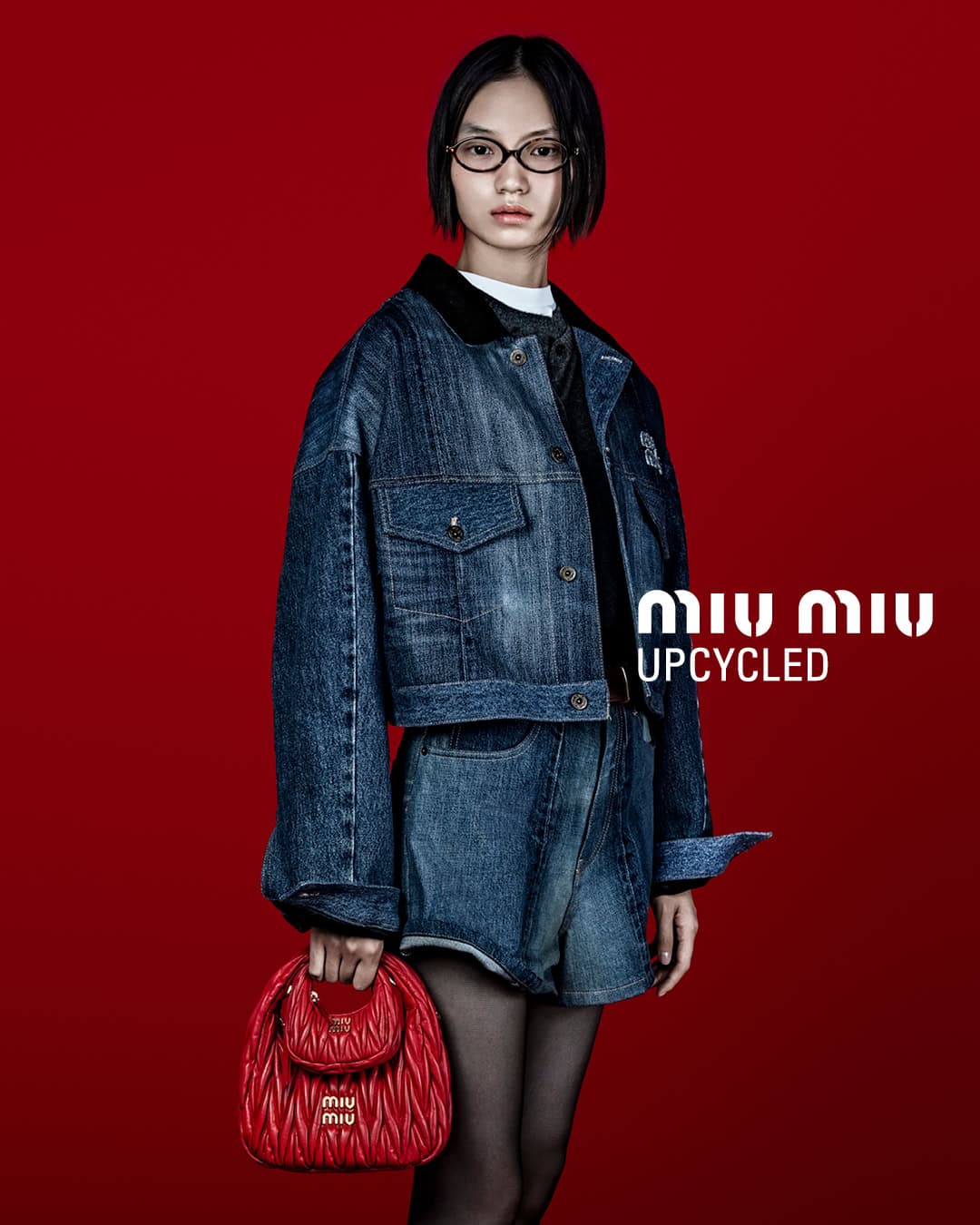 Fashion shot of an Asian woman wearing a denim jacket and shorts, holding a red quilted Miu Miu purse against a red backdrop.
