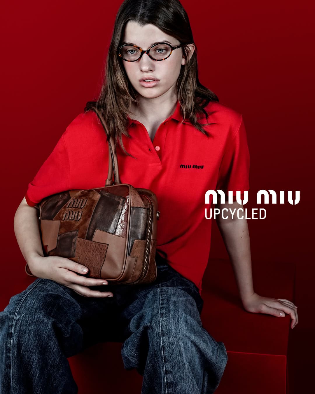Young woman in red polo and jeans sits holding brown patchwork Miu Miu bag against red backdrop, wearing tortoise-shell glasses.