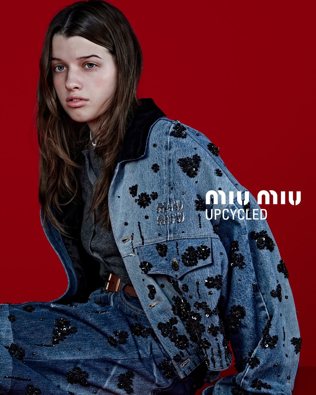 A model wears a blue denim jacket and jeans with black embellishments against a red backdrop in a fashion advertisement.