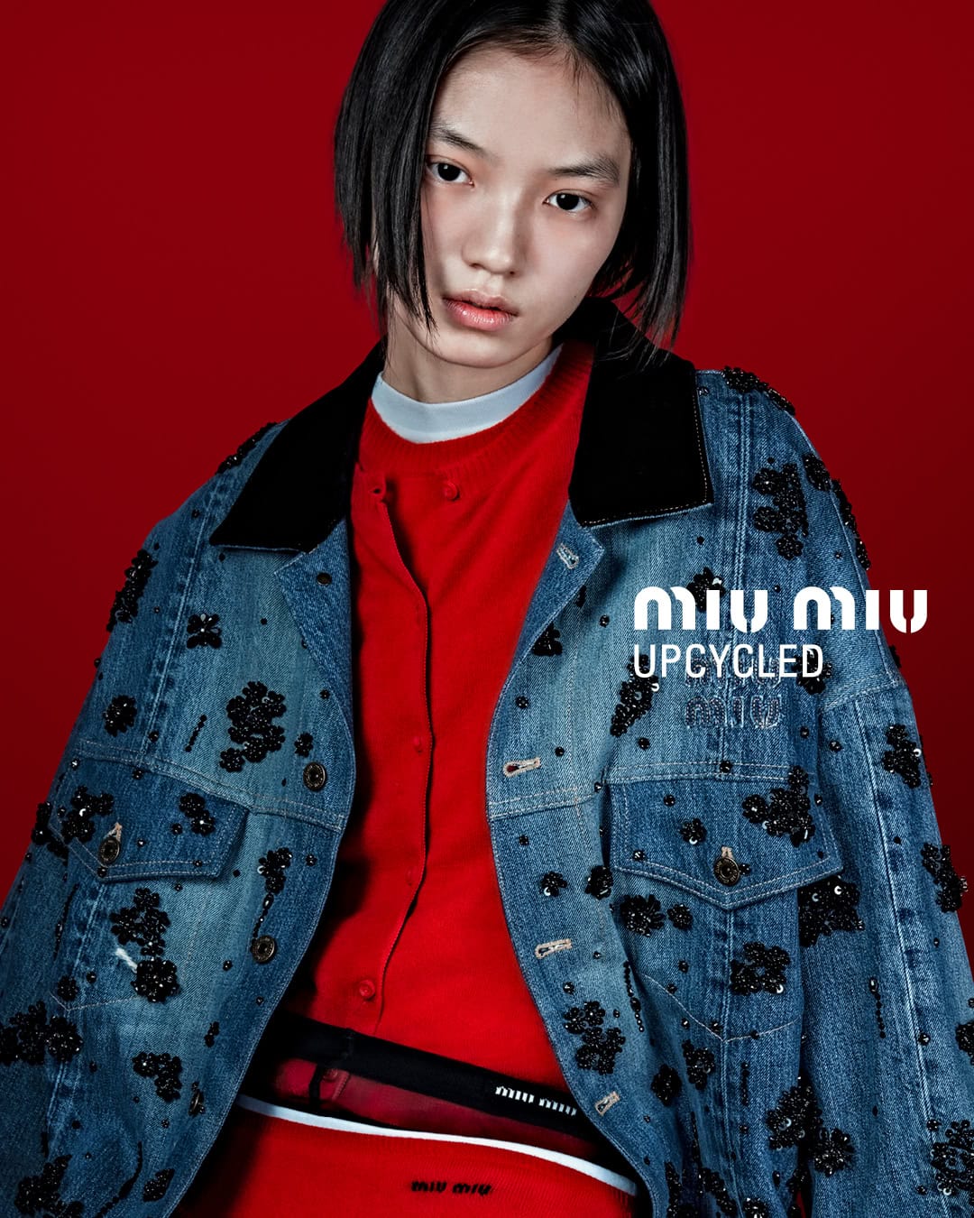 Asian female model wearing a decorated denim jacket and red outfit against a red backdrop, showcasing modern fashion style.