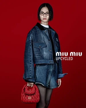 Model in denim jacket and shorts holding a red handbag against a solid red studio background for Miu Miu