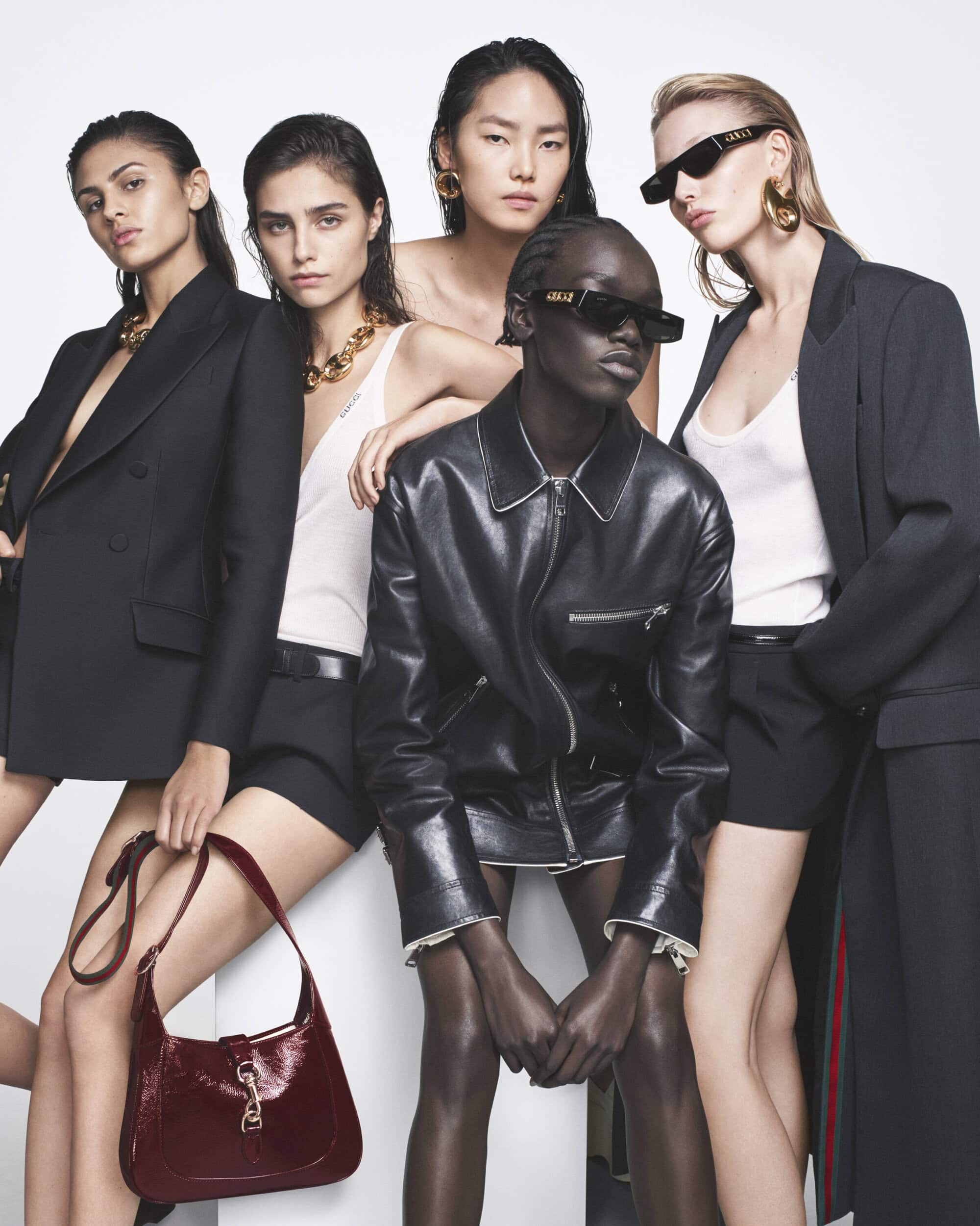 Five women pose in a studio wearing contemporary jackets, tops, and bottoms. One woman holds a maroon handbag. Sunglasses and jewelry visible.