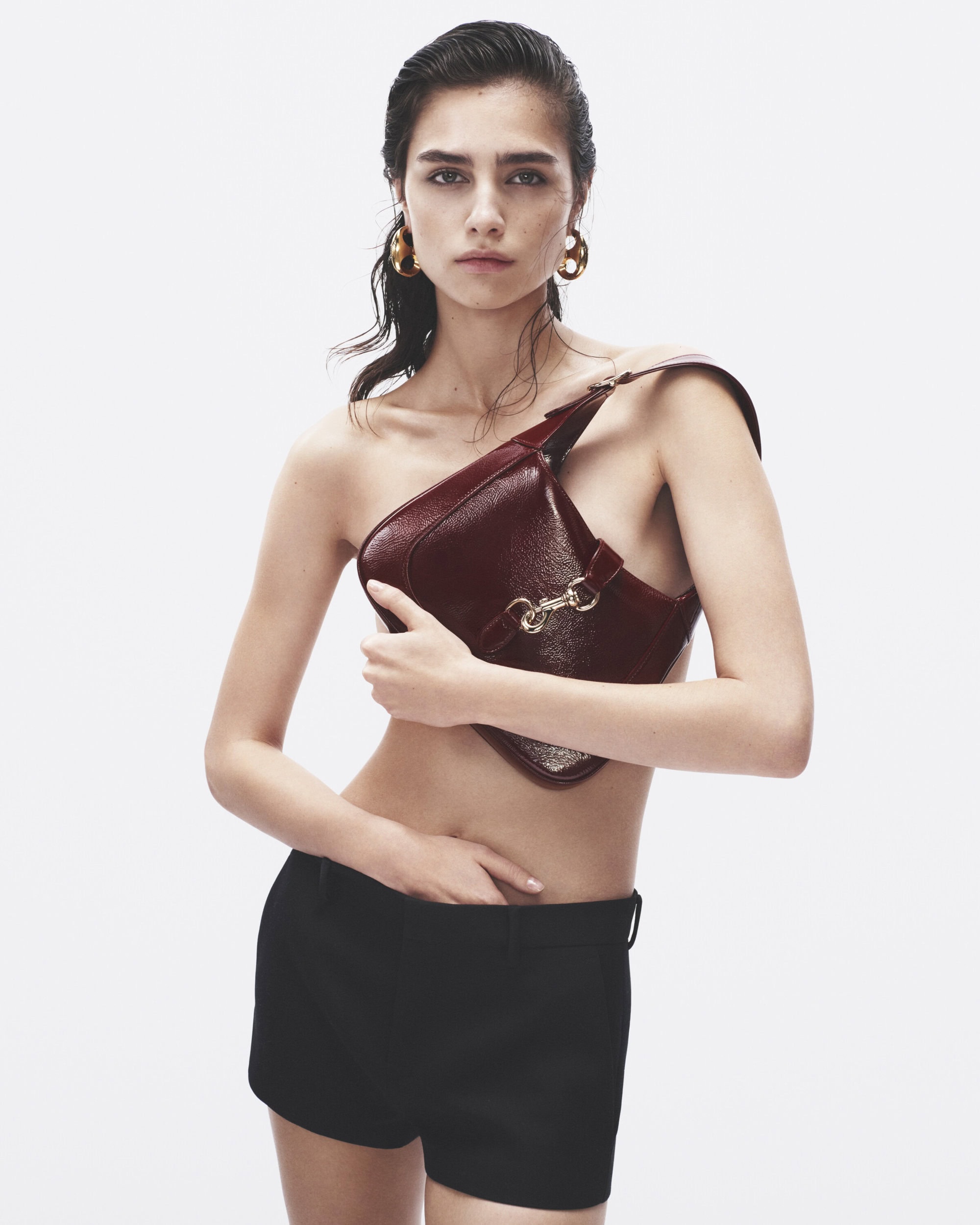 Fashion shot of a woman with dark hair and gold earrings holding a shiny, dark red purse in a bright, white studio setting.
