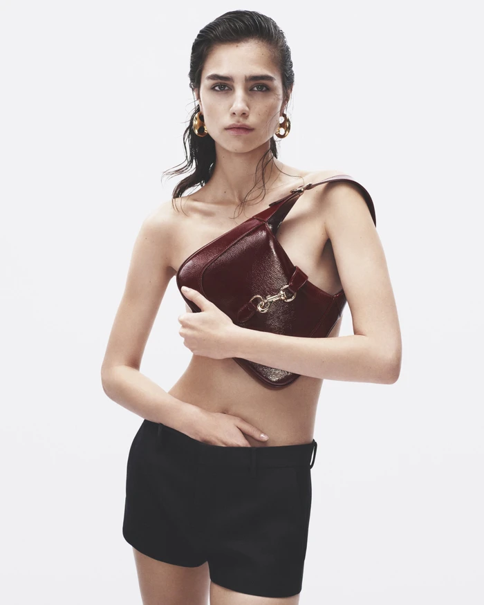 Model wearing dark shorts holding a burgundy leather handbag against a plain white background