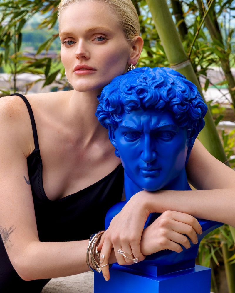 Portrait of a woman with blonde hair, wearing jewelry, posing next to a blue bust outdoors against a green bamboo backdrop.