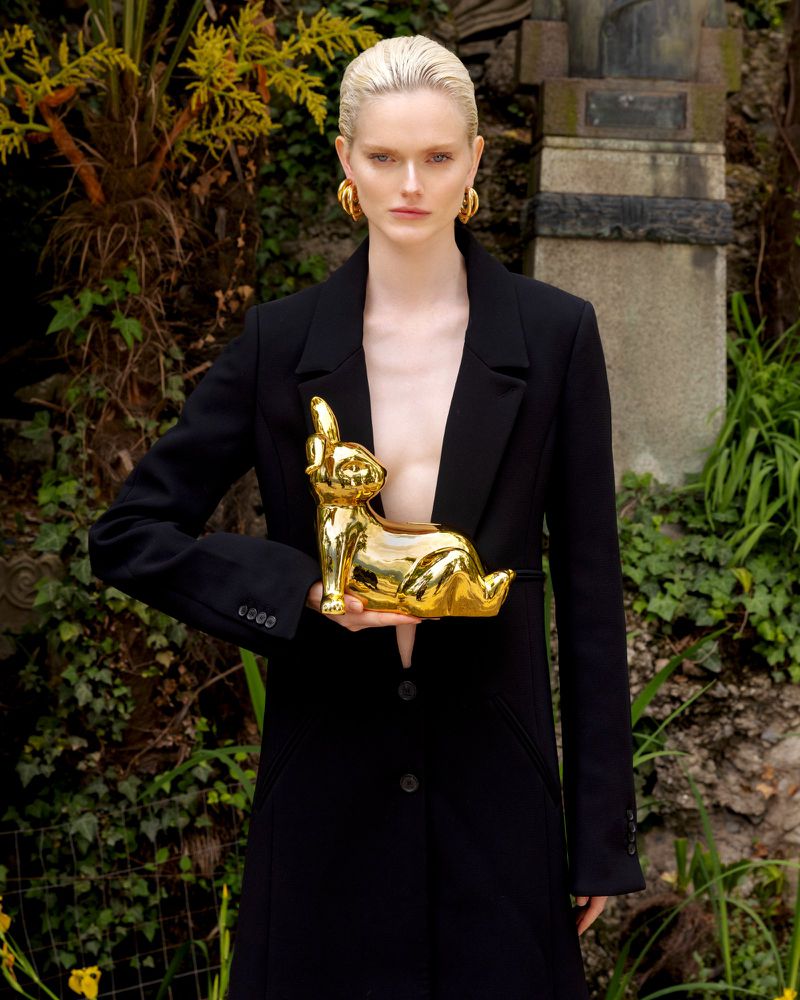 A blonde woman in a black blazer holds a golden bunny statue in an outdoor setting with greenery and stone architecture in the background.