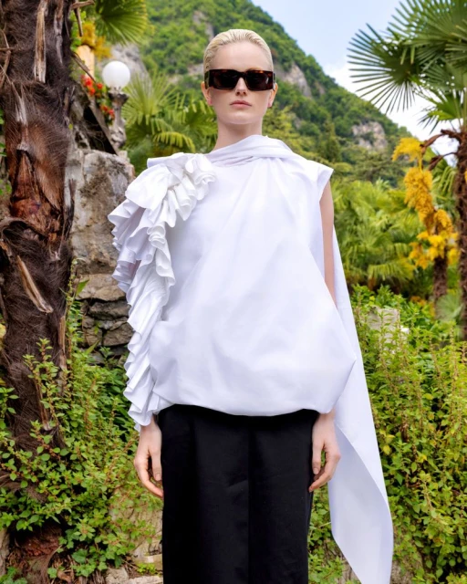 Fashionable adult female model poses outdoors wearing a white sculptural top and black pants against lush greenery and a mountain backdrop.