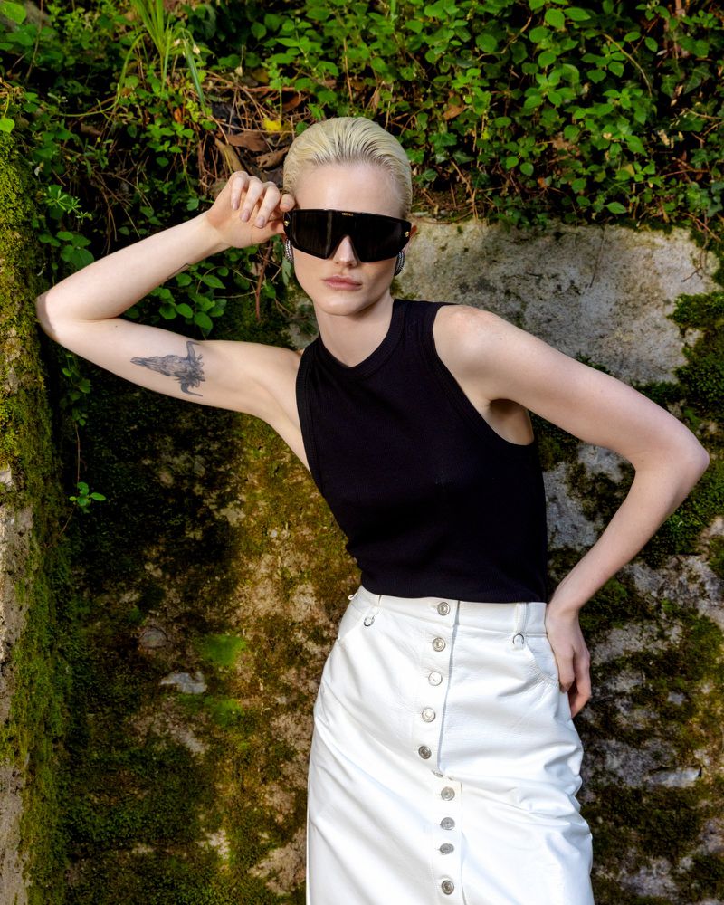 A light-skinned woman with platinum blonde hair poses in sunglasses, a black top, and a white buttoned skirt against a mossy wall.