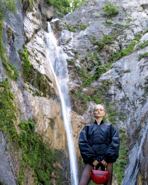 Woman in black jacket poses with a red handbag in front of a waterfall cascading down a rocky cliff covered with green vegetation.