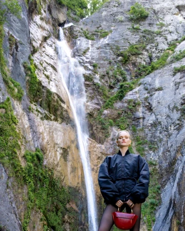 Woman in black jacket poses with a red handbag in front of a waterfall cascading down a rocky cliff covered with green vegetation.