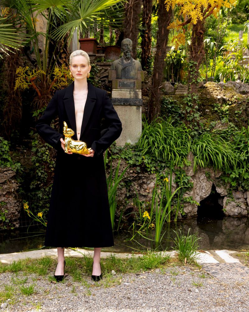 Woman in black coat holds gold rabbit statue in lush green garden with stone bust in background, fashion editorial photography.