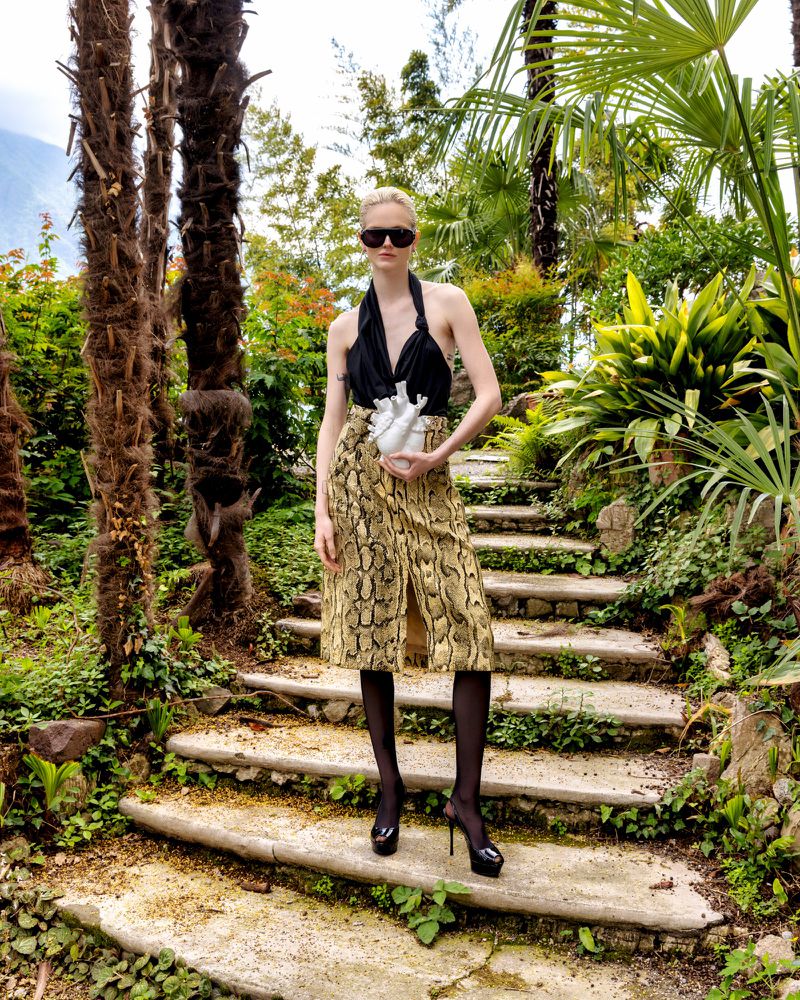 A fair-skinned fashion model on stairs with plants wearing a black top, snakeskin print skirt, dark tights, and platform high heels.