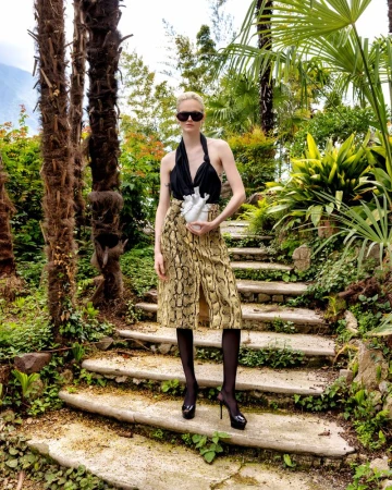 A fair-skinned fashion model on stairs with plants wearing a black top, snakeskin print skirt, dark tights, and platform high heels.