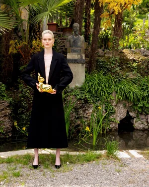 Model standing outdoors wearing a long black coat and holding a golden rabbit figurine near a pond and stone garden statue.