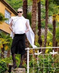 Model wearing a white ruffled top and black skirt posing outdoors near palm trees for Elle Lithuania