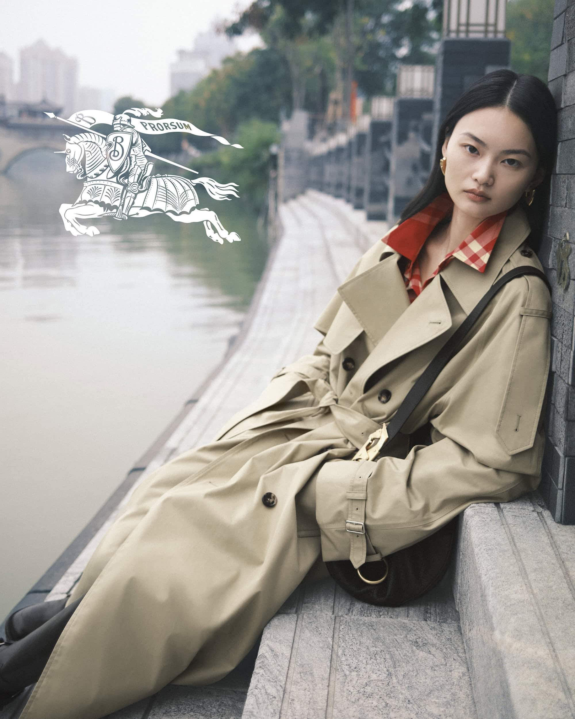 Asian woman in a trench coat leans against a wall next to a river in an urban scene with a logo in the background.