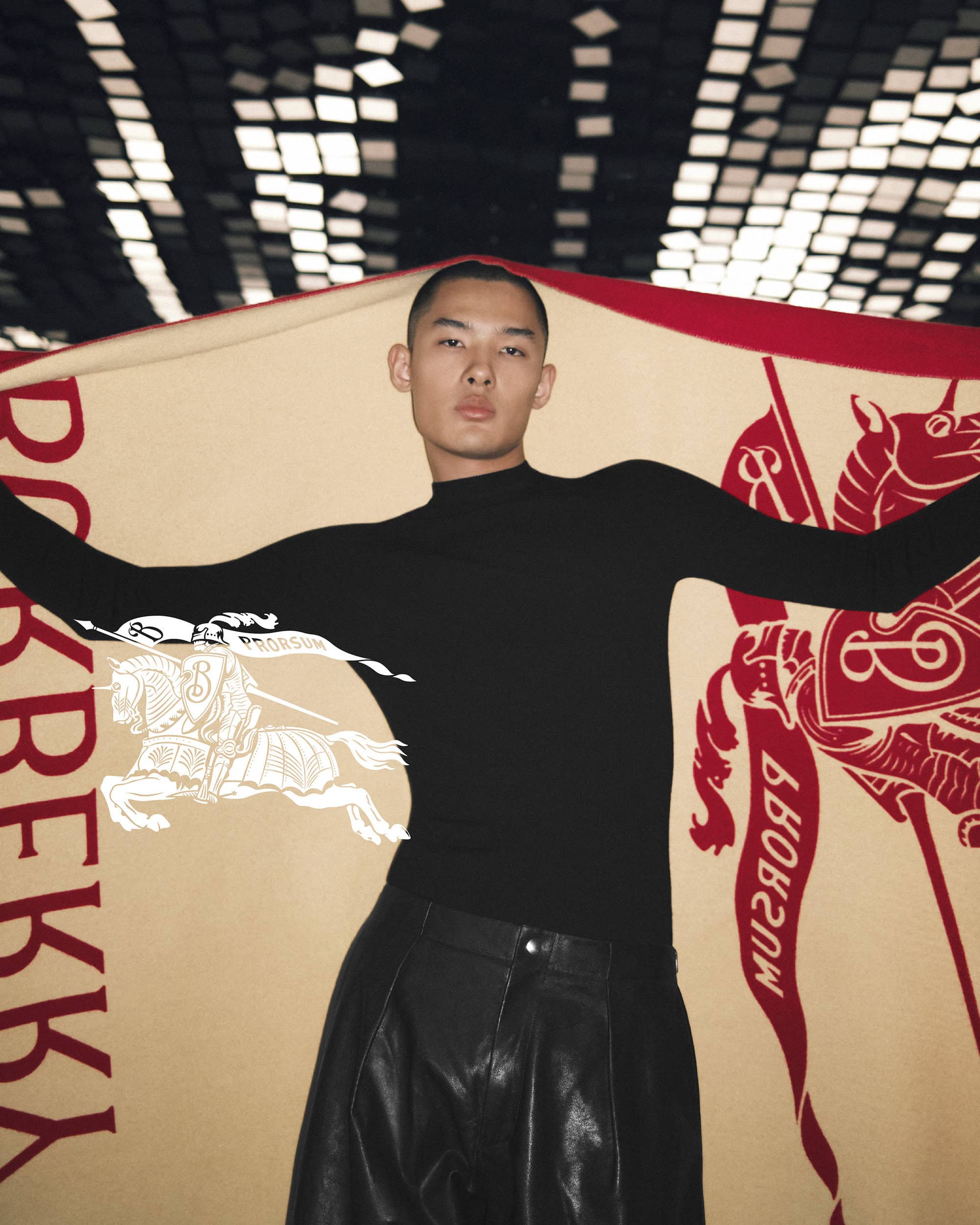 An adult Asian male poses indoors with a Burberry blanket featuring a horse-and-rider logo. He is wearing a black top and shiny black pants.