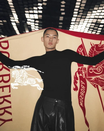 An adult Asian male poses indoors with a Burberry blanket featuring a horse-and-rider logo. He is wearing a black top and shiny black pants.