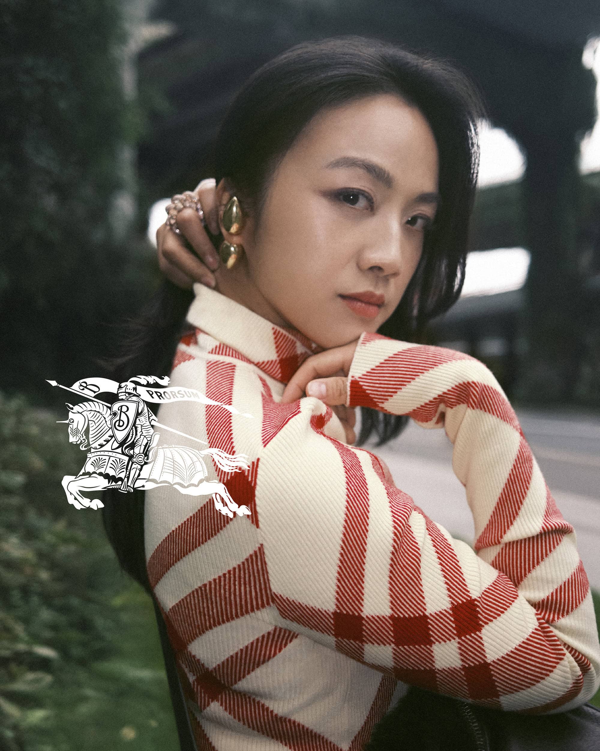 Portrait of a serene Asian woman wearing a red and white plaid sweater, accessorized with gold jewelry, posing outdoors against a leafy backdrop.