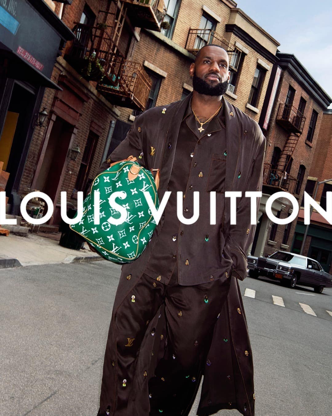 LeBron James stands on a city street with a green Louis Vuitton bag, wearing a brown patterned pajama set and gold chain. 