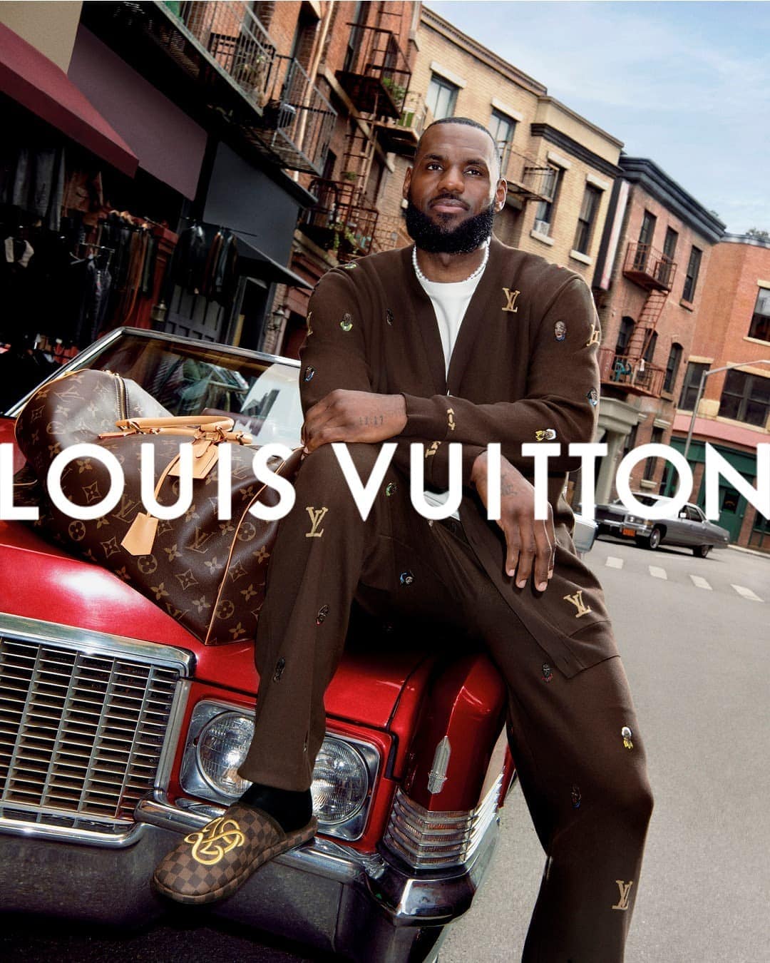 LeBron James poses on a red car with a Louis Vuitton bag in an urban setting with brick buildings in a fashion advertising campaign.